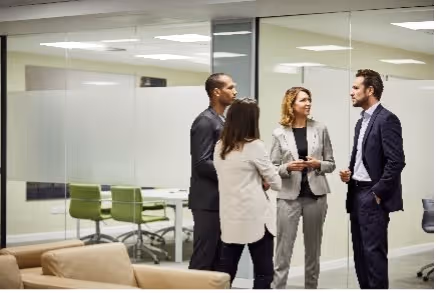 Vier mensen in zakelijke kleding praten met elkaar in een kantoorruimte met glazen wanden.