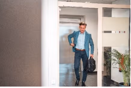 Man in een blauw colbert loopt een kantoorruimte in met een zwarte aktetas in de hand.