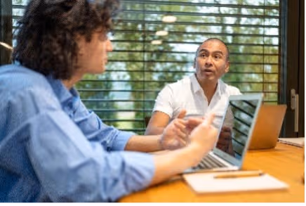 Twee mensen praten aan een tafel met laptops; de man luistert aandachtig naar de ander.