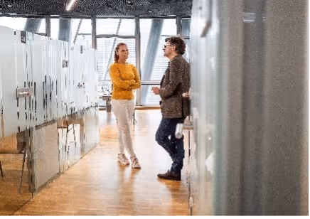 Man en vrouw praten met elkaar in een gang met glazen wanden en houten vloer.