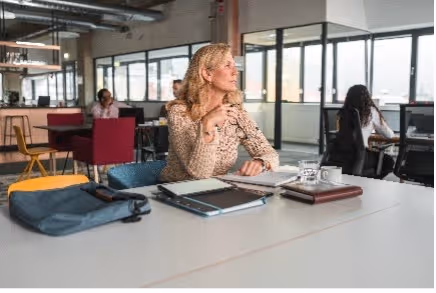 Vrouw met bril zit aan een bureau in een open kantoor en kijkt opzij met een pen in haar hand.