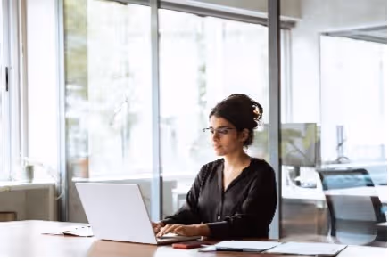 Vrouw werkt geconcentreerd op een laptop aan een bureau in een lichte ruimte met glazen wanden.
