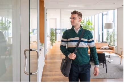Man met bril en groene trui loopt een modern kantoor binnen met houten vloer en veel daglicht.