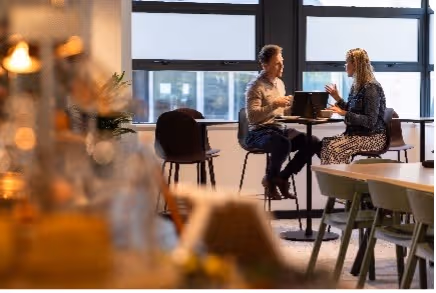 Twee mensen zitten aan een hoge tafel in gesprek met een open laptop tussen hen in.