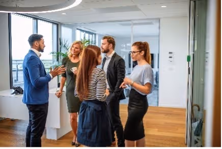 Vijf collega’s staan bij elkaar in gesprek in een modern kantoor met houten vloer.