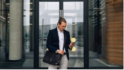 Man in een donker jasje kijkt op zijn telefoon terwijl hij buiten voor een glazen ingang staat.