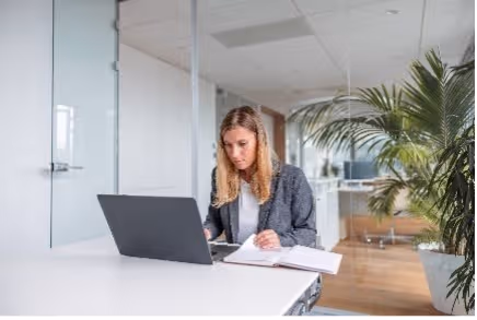 Vrouw werkt achter een laptop aan een wit bureau in een lichte kantoorruimte met planten.