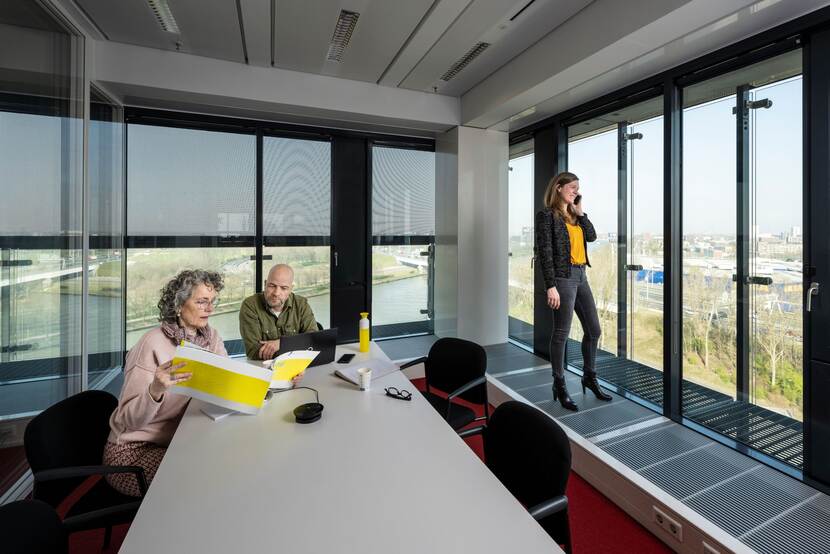 Twee mensen overleggen aan tafel, vrouw telefoneert staand er naast